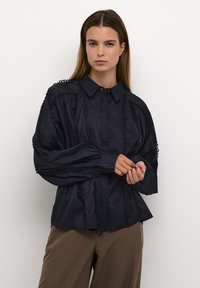 Schwarze Bluse mit Knopfverschluss und Puffärmeln, strukturierte Details entlang der Nähte und glatte Oberfläche. Kombiniert mit braunen, locker sitzenden Hosen.