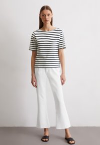 Gestreiftes Kurzarmoberteil in Weiß und Marineblau mit einem Bootsausschnitt. Kombiniert mit weißen Schlaghosen und schwarzen Rutsch-Sandalen.