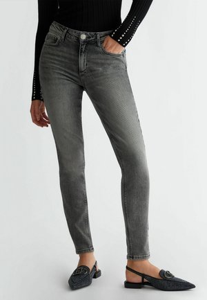 Jean noir délavé slim porté avec des ballerines slingback noires à clous et un haut noir à manches longues avec des poignets cloutés. Une main dans la poche.