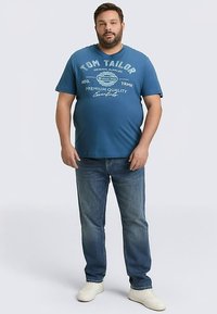 Blauw katoenen T-shirt met witte tekst en grafische accenten, gecombineerd met lichtblauwe spijkerbroek en witte sneakers, staand tegen een effen achtergrond.