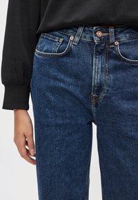 Záber zblízka na stred tela v tmavomodrých rifliach s kontrastným prešitím a meďovými nitmi, doplnených o čierne tričko s dlhým rukávom.