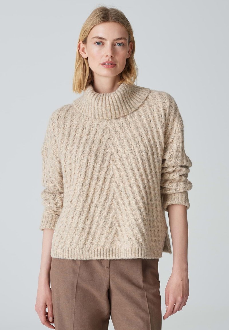 Beiger, strukturierter Strickpullover mit hohem, geripptem Kragen und diagonalen Mustern. Weite Ärmel und Saum, kombiniert mit braunen Hosen.