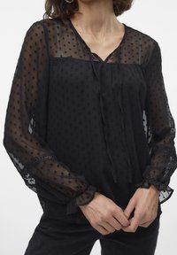 Blouse noire transparente à texture à pois, encolure en V, détail noué, et manches longues avec poignets volantés. Le tissu est léger et aéré.
