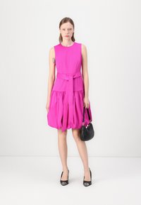 Fuchsiové šaty bez rukávů s uvázaným pasem, sklady v sukni a hladkým povrchem; doplněné malou černou kabelkou a černými lodičkami.