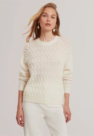 Kvinde med langt blondt hår iført perleøreringe med dråbeform, en cremefarvet kabelstrikket sweater og hvide bukser mod en ensfarvet baggrund.