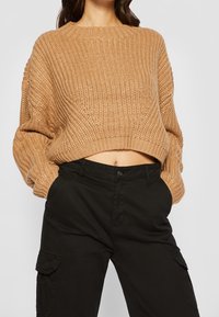 Beskuren, stickad tröja i mjuk beige med ett strukturerat mönster. Har en vid halsringning och lösa ärmar, kombinerad med svarta cargobyxor.