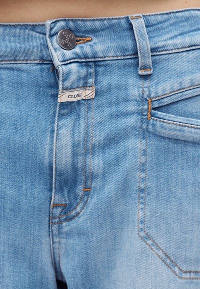 Κοντινό πλάνο στη μέση τζιν από ανοιχτό μπλε denim με μεταλλικό κουμπί και μικρή υφασμάτινη ετικέτα που γράφει "CLOSED."
