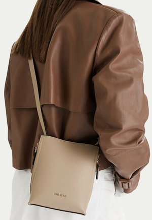 Osoba nosící hnědou koženou bundu a bílé kalhoty má přes rameno béžovou kabelku Gino Rossi.