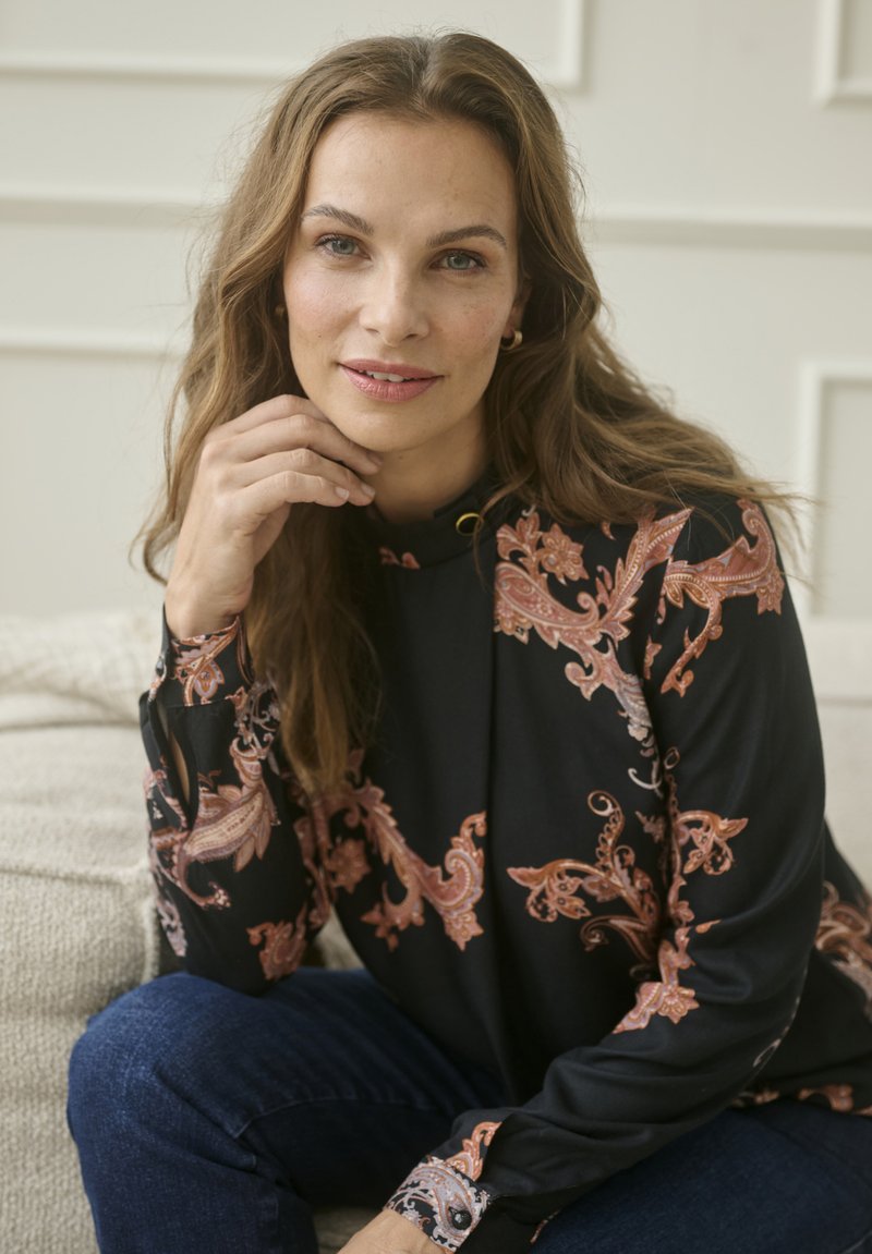 Hosszú hajú nő fekete felsőt visel rózsaszín paisley mintákkal, egy kanapén ülve, állát a kezére támasztja, és a kamerába néz.