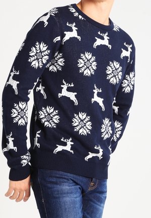 Pull bleu marine avec motifs blancs de rennes et de flocons de neige porté par une personne habillée d’un jean bleu.