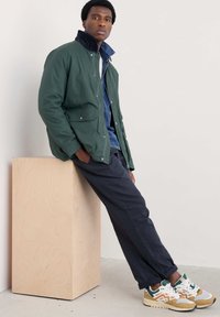 Chaqueta de lona verde con cuello negro, combinada con una camisa de mezclilla, pantalones oscuros y zapatillas de varios colores. Diseño sencillo, con bolsillos funcionales.