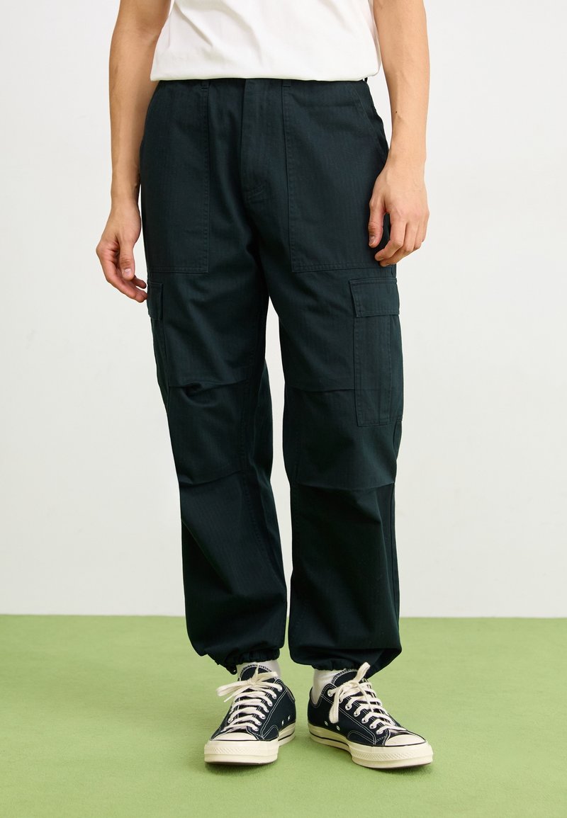 Personne portant un pantalon cargo noir avec des poignets élastiques, des chaussettes blanches, des baskets en toile noires et un t-shirt blanc, debout sur un sol vert.