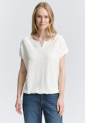 Haut blanc à manches courtes avec une finition texturée, décolleté fendu et coupe loose, assorti à un jean bleu foncé.