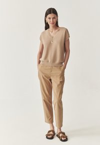 Vrouw die met haar handen in de zakken staat, gekleed in een beige gebreid mouwloos topje, kaki broek, bruine sandalen en gouden sieraden tegen een witte achtergrond.