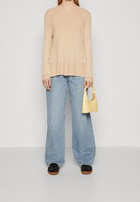 Pull beige en maille à manches longues, jean bleu clair à jambes larges, chaussures noires, tenant un petit sac à main jaune avec une poignée incurvée.