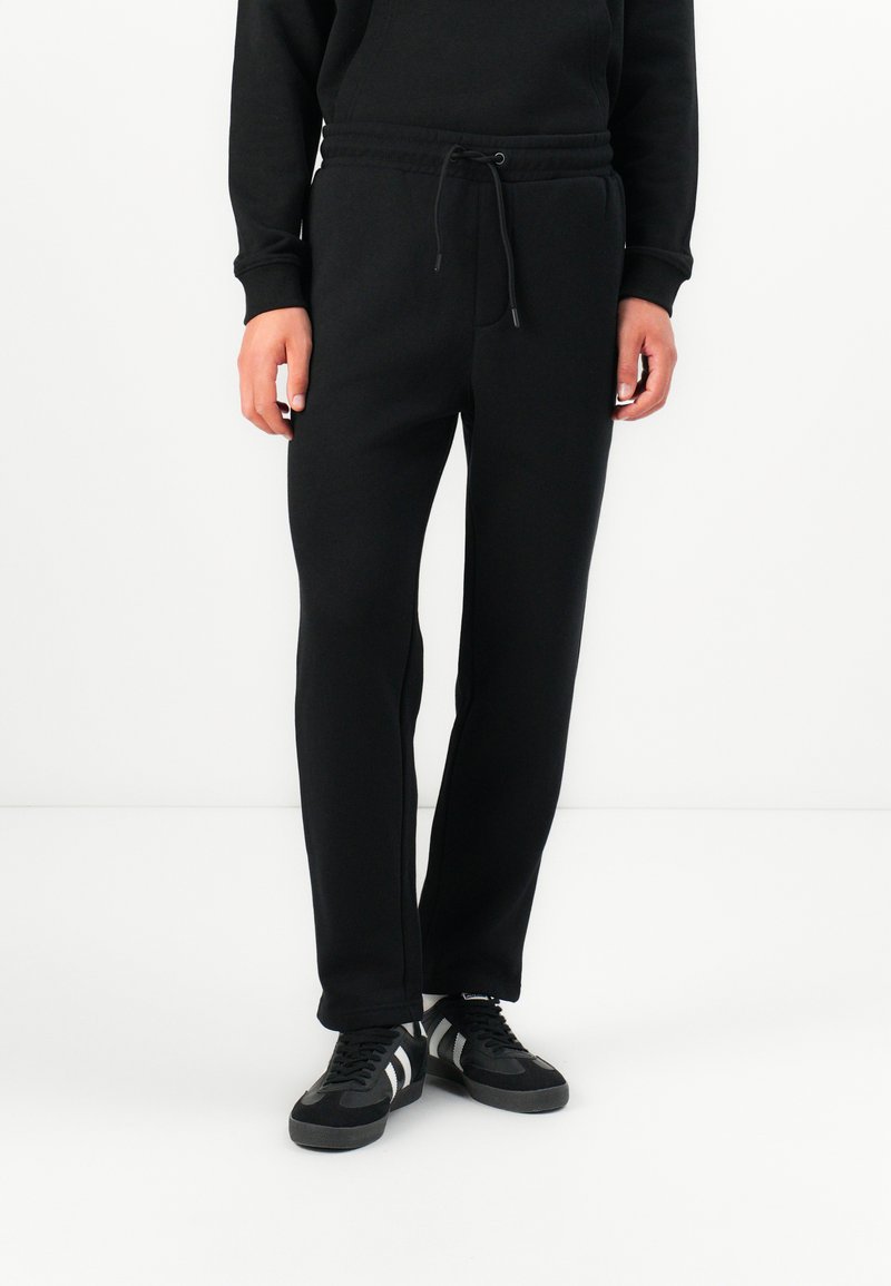 Black sweatpants made of cotton with a drawstring waist, tapered legs, and side pockets. Worn with black sneakers featuring white stripes.