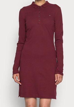 Nainen pukeutuneena pitkähihaiseen, burgundinväriseen poolomekkoon, jossa on napillinen läppä ja pieni logo rinnassa, seisoo yksiväristä taustaa vasten.