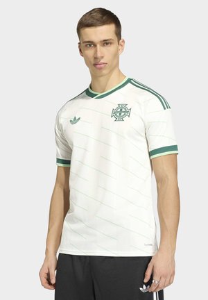 Modèle masculin portant un maillot de football d'Irlande du Nord blanc et vert avec le logo Adidas et un short noir, sur un fond clair uni.