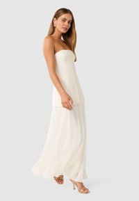 Trägerloses weißes Kleid mit tailliertem Oberteil und fließendem, transparentem Rock. Goldfarbene High-Heelsandalen und auffällige Ohrringe vervollständigen den Look.