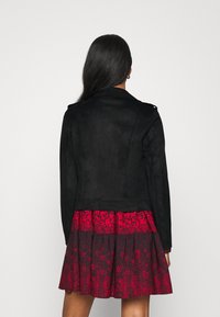 Giacca nera in suede con colletto strutturato e spalline, abbinata a un vestito corto rosso a fiori con pois neri.