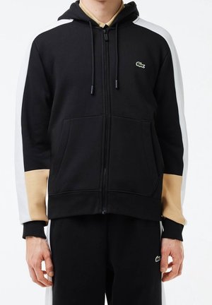 Person trägt einen schwarzen Hoodie mit Reißverschluss, braunen und weißen Ärmeln sowie passende schwarze Hosen, auf beiden mit sichtbarem Krokodil-Logo.