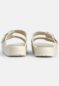 Sandales beiges à enfiler avec trois larges brides et des accents floraux. Fabriquées en matériau flexible avec des semelles texturées pour une meilleure adhérence.