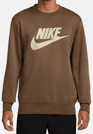 Homme portant un sweat-shirt marron à manches longues avec un grand logo blanc Nike et un swoosh sur la poitrine, associé à un pantalon noir.
