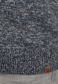 Suéter de punto grueso en color navy con motas blancas, dobladillo acanalado y parche de logo de cuero marrón en la parte inferior. Textura suave, diseño de trenzado.