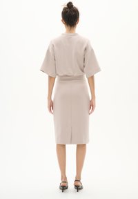 Beiges Kleid mit taillierter Silhouette, kurzen weiten Ärmeln und einem Rückenschlitz. Glattes Material, knielang, kombiniert mit schwarzen Absatzsandalen.