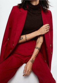 Červený blazer a odpovídající kalhoty, přiléhavý střih, hladká textura. Hnědý rolák pod ním, který ukazuje propracované tetování na rukou.