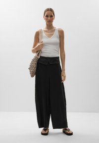 Débardeur blanc à côtes et pantalon noir à jambe large avec une ceinture repliée. Sac tissé marron. Le modèle se tient contre un fond neutre.