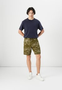 Navy blue t-shirt and olive green shorts with white floral embroidery. Model wearing white sneakers; minimalist background.