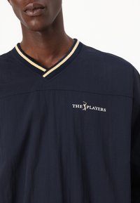 Tmavomodrá polo košeľa s výstrihom do V a bielymi a žltými pruhmi. Vyšitý logo "THE PLAYERS" nad grafikou golfistu.