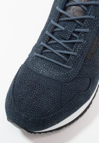 Granatowy sneaker z chropowatą cholewką z zamszu, posiadający perforacje, płaskie sznurowadła oraz białą środkową podeszwę. Czarna podeszwa zewnętrzna z wzorem do przyczepności.