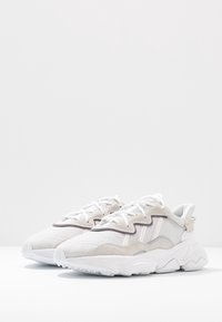 Pár světle béžových a bílých sportovních tenisek s vlnitými semišovými akcenty a silnými texturovanými podrážkami na bílém povrchu.