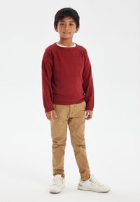 Maglione rosso a maglia con polsini a costine, pantaloni beige e sneakers bianche. Il design presenta un collo rotondo e una superficie texturizzata. Dito sulla vita.