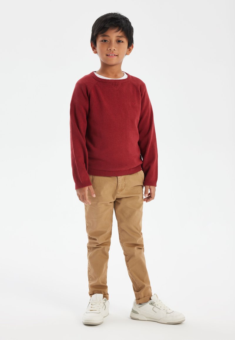 Maglione rosso a maglia con polsini a costine, pantaloni beige e sneakers bianche. Il design presenta un collo rotondo e una superficie texturizzata. Dito sulla vita.