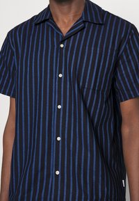 Camicia a maniche corte con strisce verticali nere e blu, colletto a punta e taschino sul petto. I bottoni sono di colore crema.