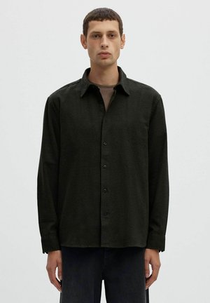 Un hombre mira a la cámara y lleva una camisa oscura abotonada sobre una camiseta marrón y pantalones oscuros, delante de un fondo liso y claro.