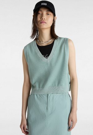 Pull sans manches vert menthe avec un col en V et un ourlet côtelé, porté sur un t-shirt noir. Associé à une jupe vert clair et une casquette noire.