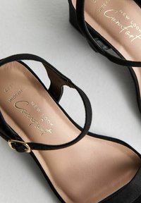 Sandalia cuña negra con correa ajustable en el tobillo, plantilla suave en color beige y detalles en dorado. Diseño de ancho cómodo.