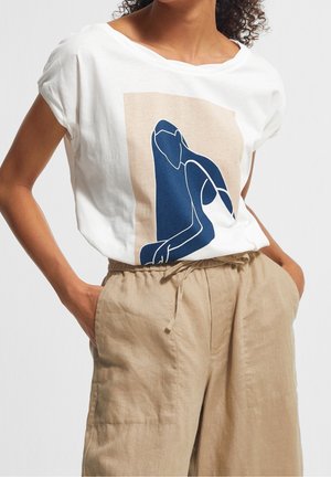 Vrouw draagt wit T-shirt met abstracte blauwe figuurprint en beige linnen broek met hoge taille, handen in de zakken.