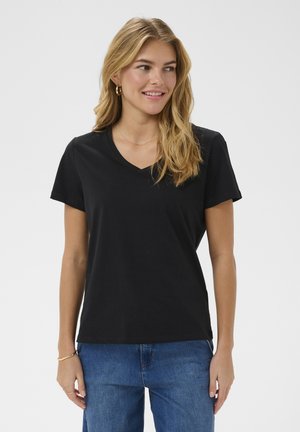 Blondínka so zlatými kruhovými náušnicami, v čiernom tričku s krátkym rukávom a véčkovým výstrihom, modrých džínsoch, usmieva sa a mierne pozerá doprava na jednoduchom pozadí.