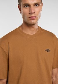 Brun bomulds t-shirt med klassisk rund hals, korte ærmer og et lille sort logo på brystet. Blød tekstur og afslappet pasform.