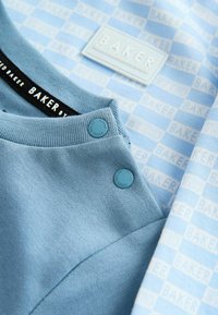 Svetlomodrý odev s textúrovanou látkou, okrúhlymi modrými cvokmi a nášivkou s logom "BAKER" na prednej strane.