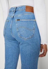 Jeans de ganga azuis de cintura alta com um patch de marca em pele, apresentando bolsos traseiros com costura amarela e um efeito de desgastado ligeiro.