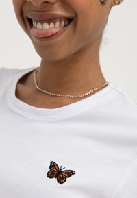 Približni kadar osobe koja se smiješi, nosi bijelu košulju s izvezenim narančastim leptirom i nježnom ogrlicom od perlica.