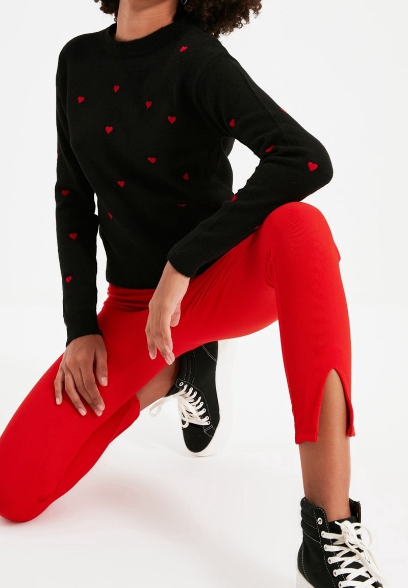 Une personne est à genoux, portant un pull noir orné de petits cœurs rouges, un pantalon rouge vif avec des fentes aux chevilles, et des baskets montantes noires avec des lacets blancs.