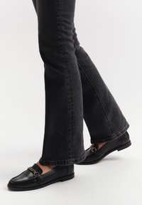 Black leather loafers with a chain detail, paired with dark flared jeans. The loafers have a smooth texture and flat sole.