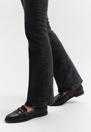Black leather loafers with a chain detail, paired with dark flared jeans. The loafers have a smooth texture and flat sole.